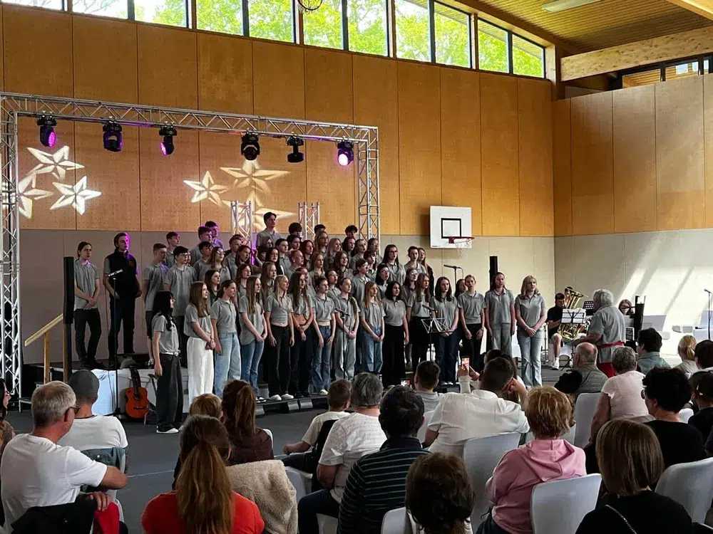 Eine große Gruppe von Jugendlichen singt auf einer Bühne vor einem Publikum in einer Halle