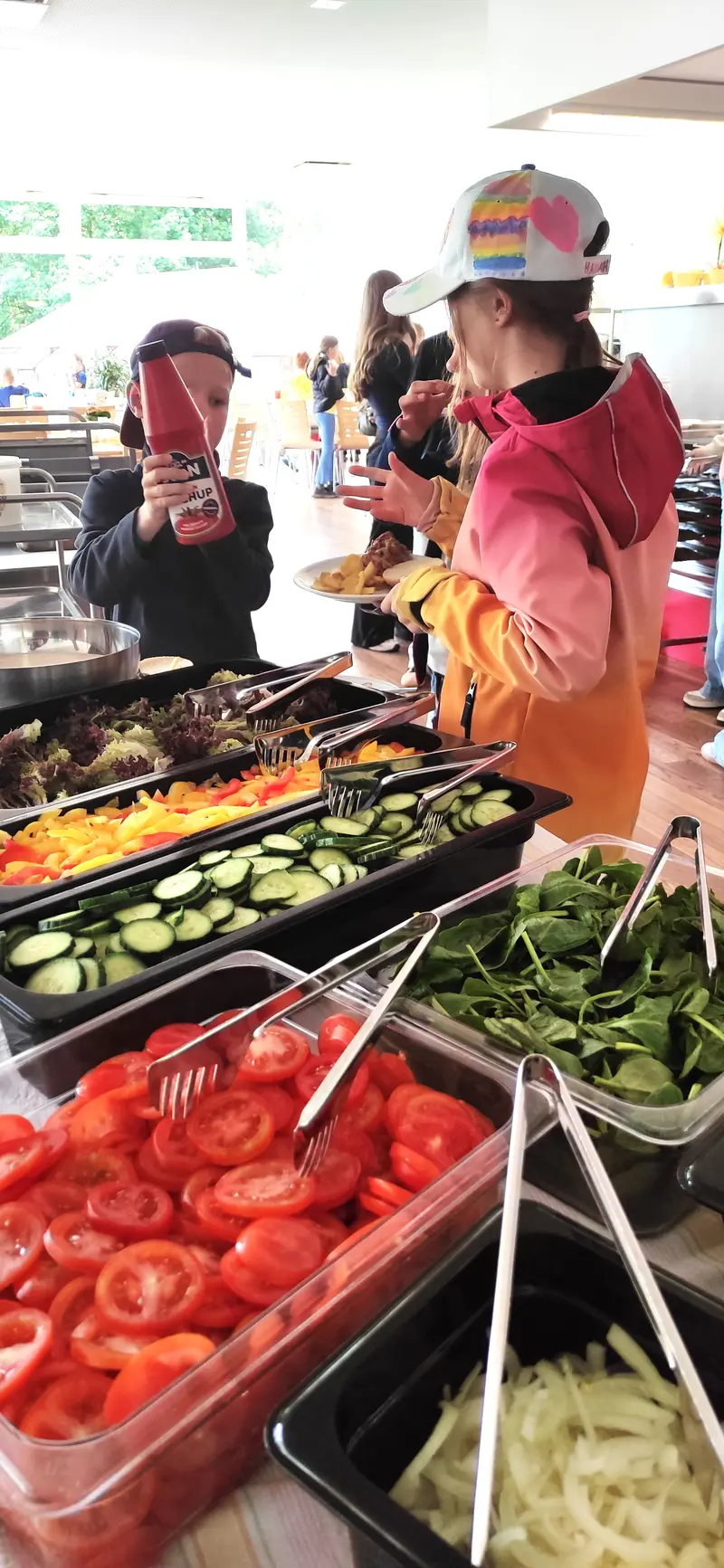 Zwei Personen essen am Buffet, lächelnd, buntes Herzcap und Gemüseauswahl