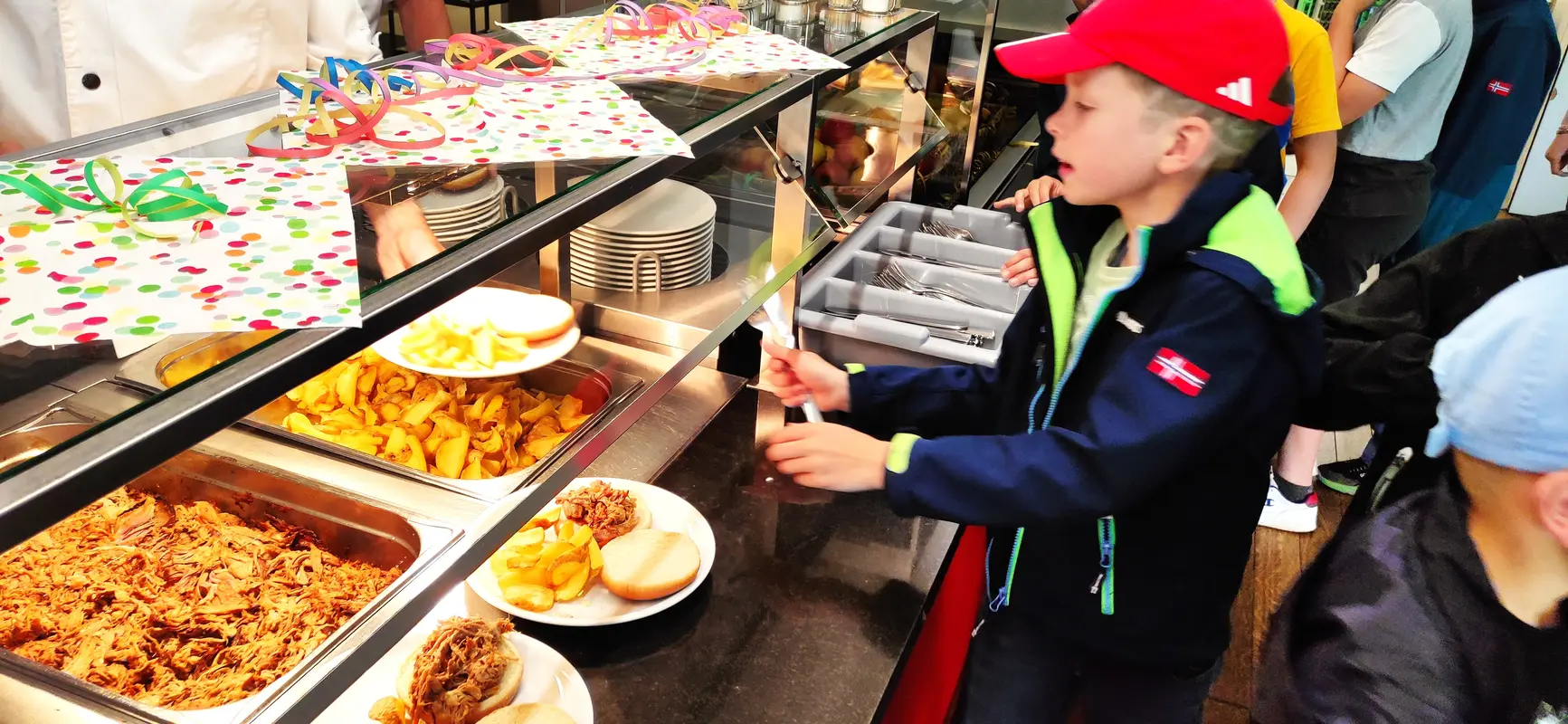 Junge mit neutralem Blick in roter Kappe am bunten Buffet mit Pommes