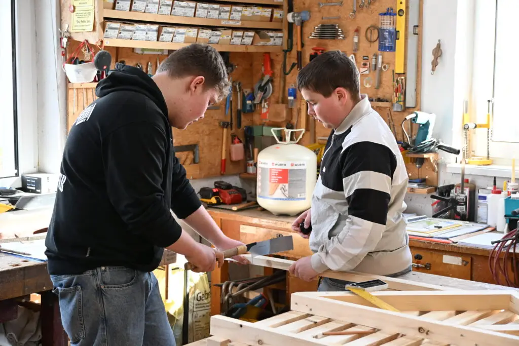 Zwei Maenner arbeiten konzentriert mit Holz in heller Werkstatt, Werkzeugwand