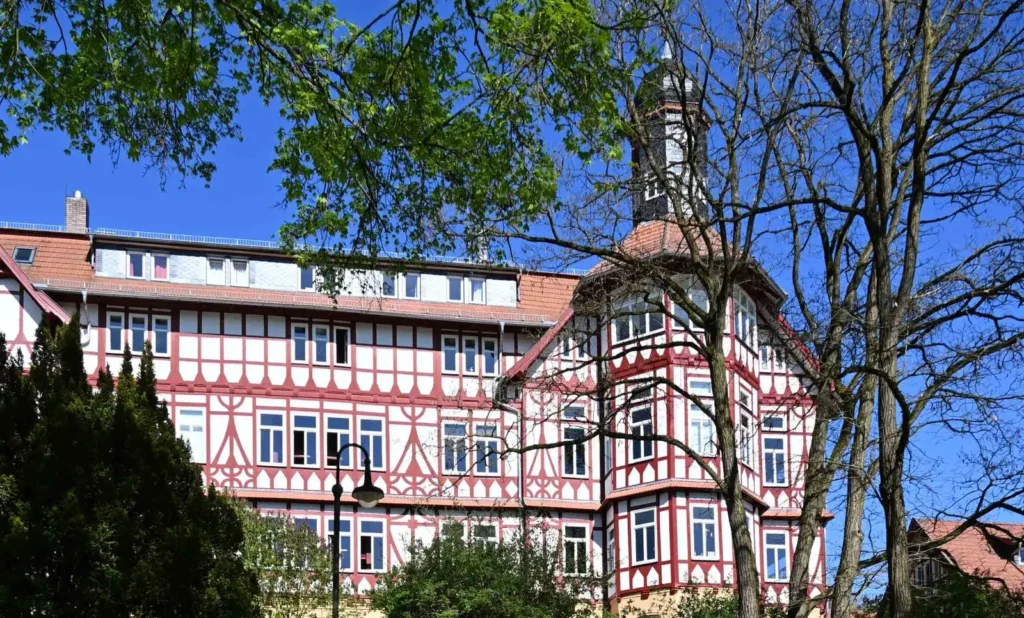 Weißes Fachwerkhaus mit roten Balken und Turm hinter Bäumen, blauer Himmel