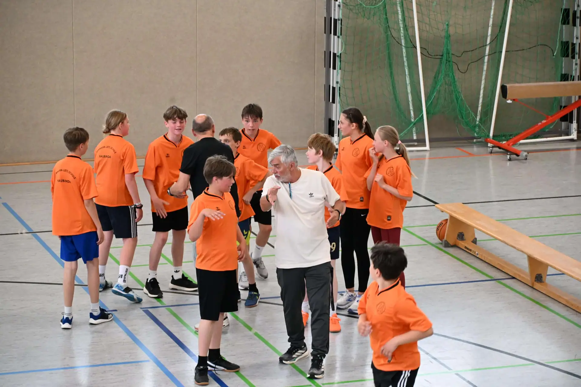 Sportgruppe in Orange lacht, Trainer in Weiß gestikuliert in Halle