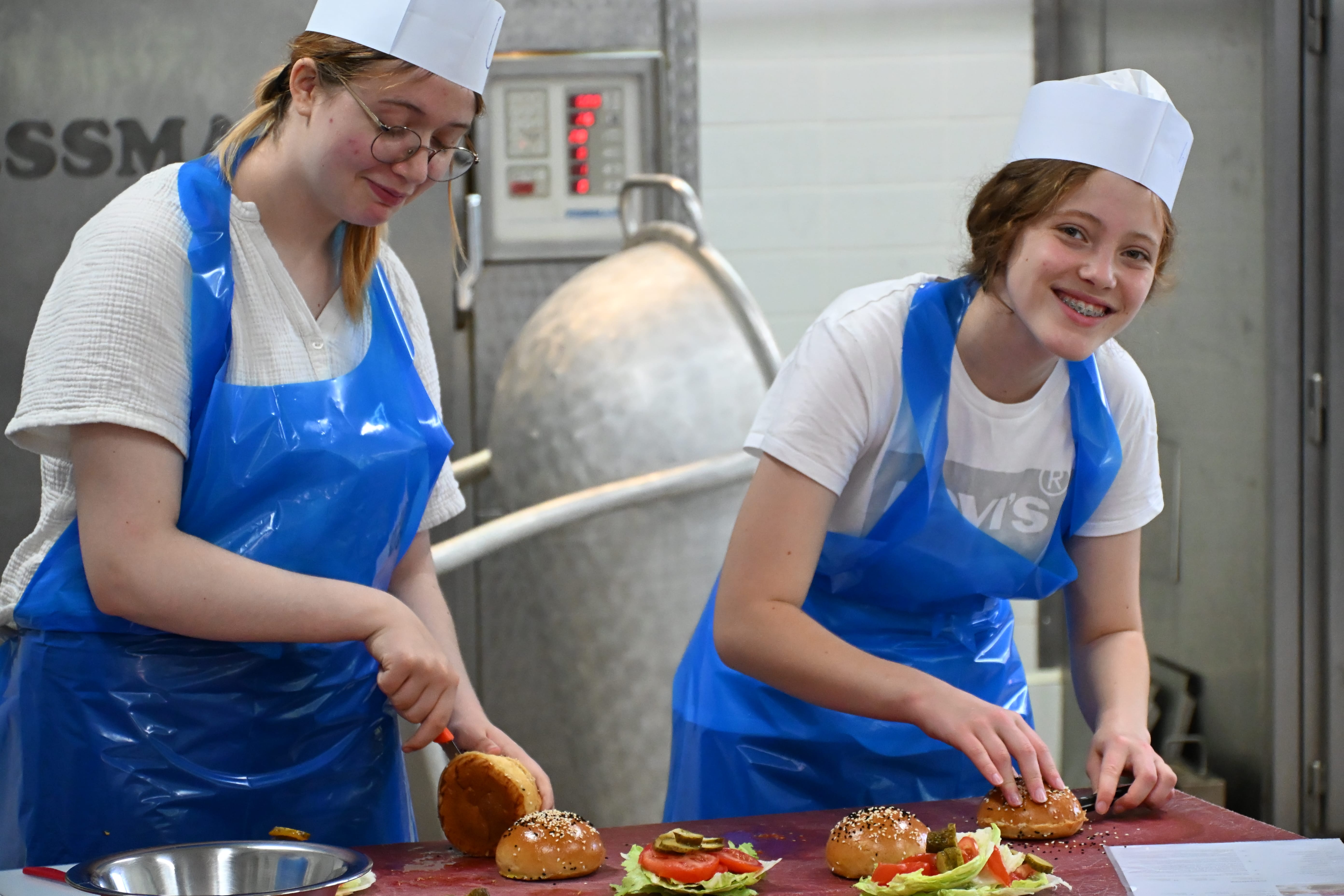 Zwei Frauen lachen beim Burgerzubereiten in blauen Schuerzen und weissen Muetzen