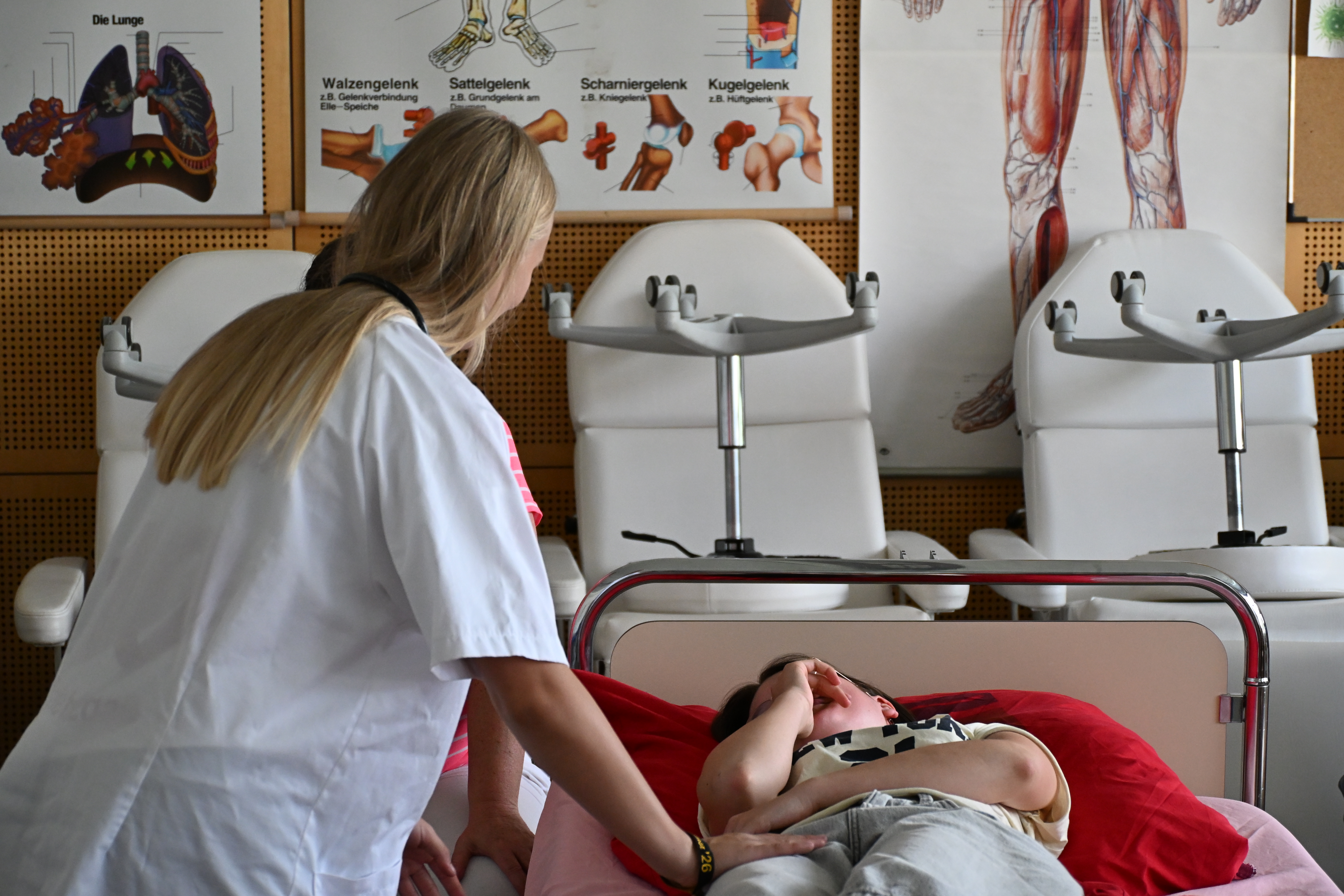 Pflegekraft ernst neben Patient auf rotem Bett in hellem Behandlungsraum