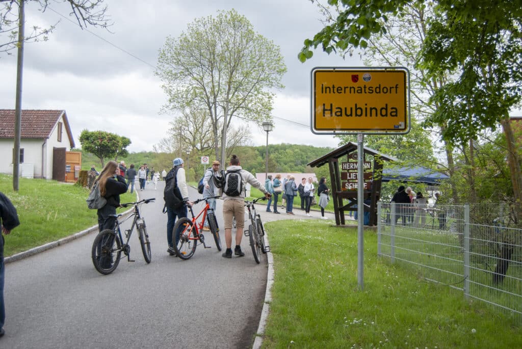 Radfahrer mit neutralem Blick neben roten und schwarzen Rädern am Ortsschild Haubinda