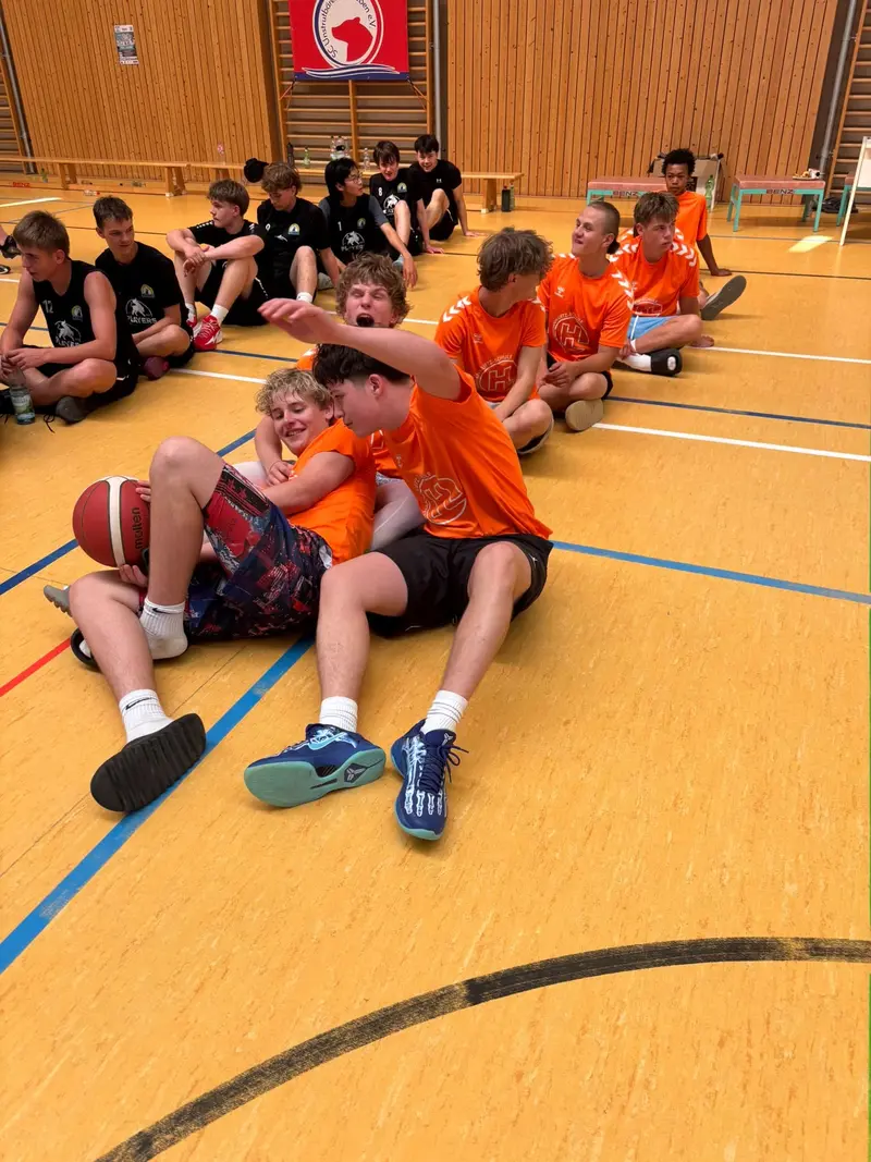 Lachende Jugendmannschaft in Orange sitzt in Halle mit Basketball