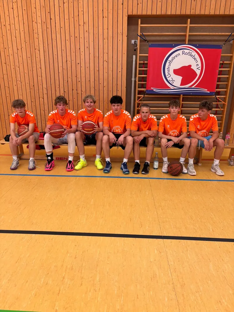 Sieben lächelnde Jungen in orangefarbenen Trikots mit Basketball in Turnhalle
