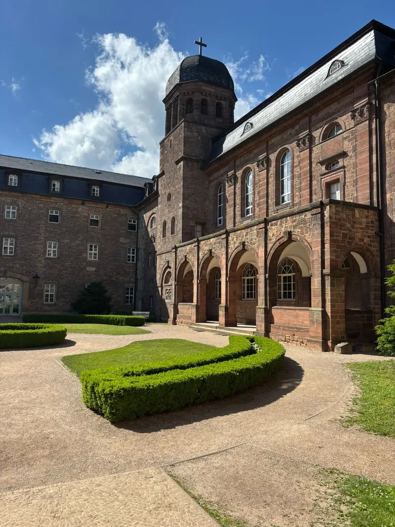 Sonniger Klosterhof mit rotem Sandsteinbau und Turm Kreuz und Bogenreihe