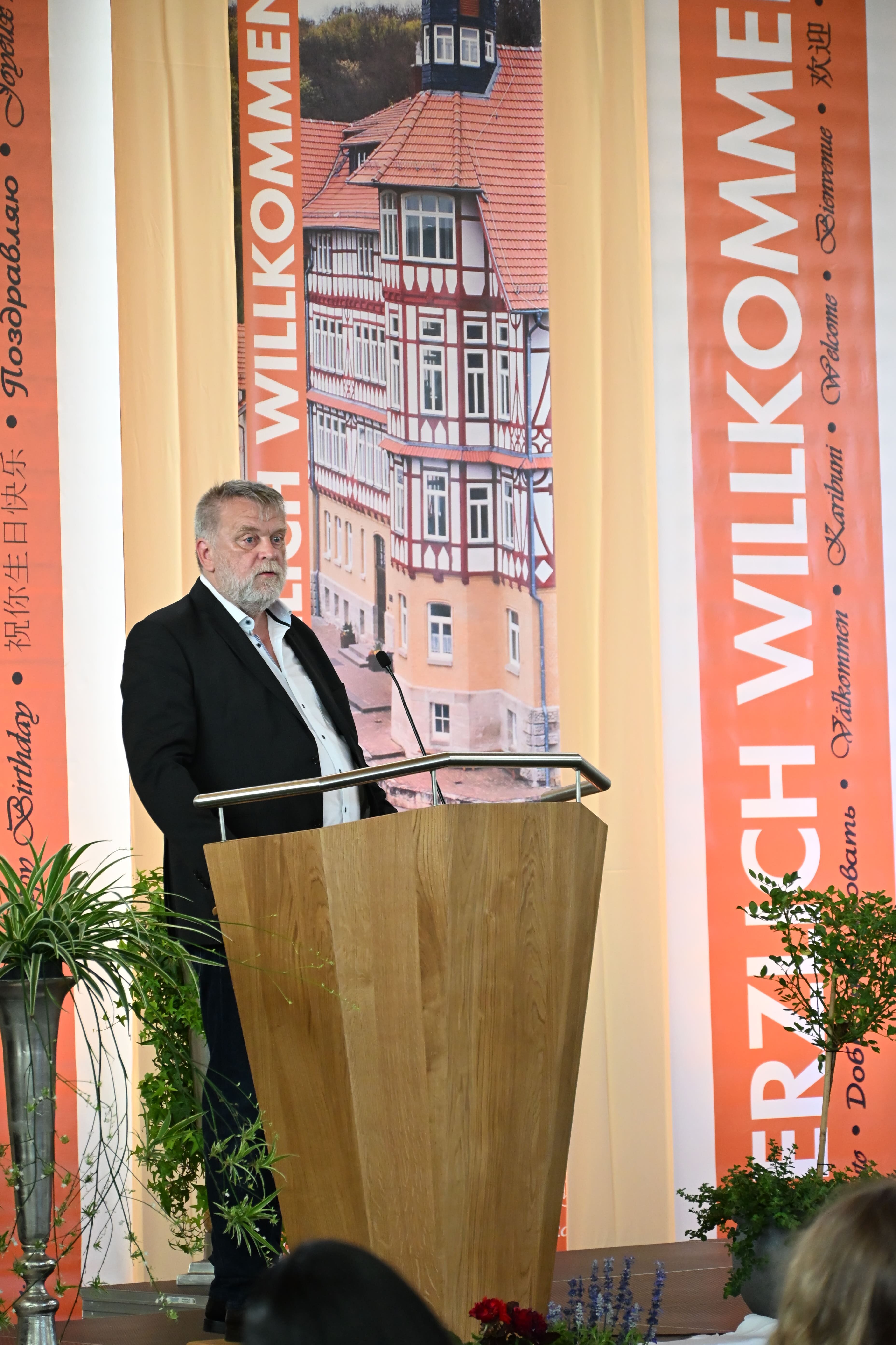 Laechelnder grauhaariger Mann im schwarzen Anzug am Holzpult vor rotem Willkommen Banner