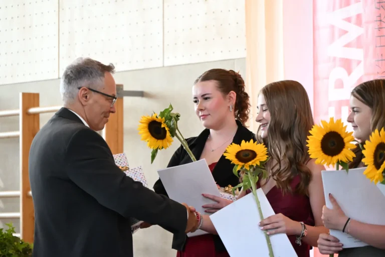 Ein Mann im Anzug gratuliert laechelnden jungen Frauen mit Sonnenblumen