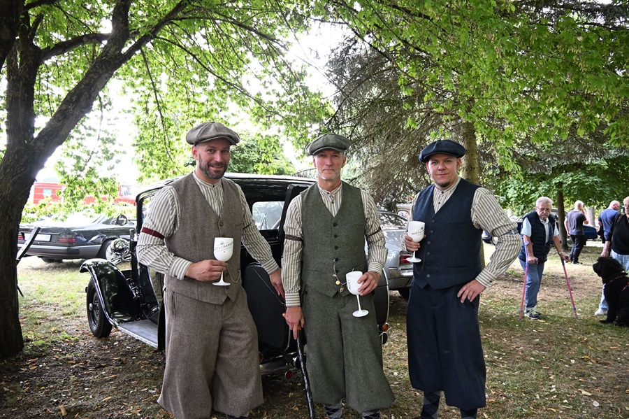Drei laechelnde Maenner in Vintage Tweed Kleidung mit Kelchen vor Oldtimer