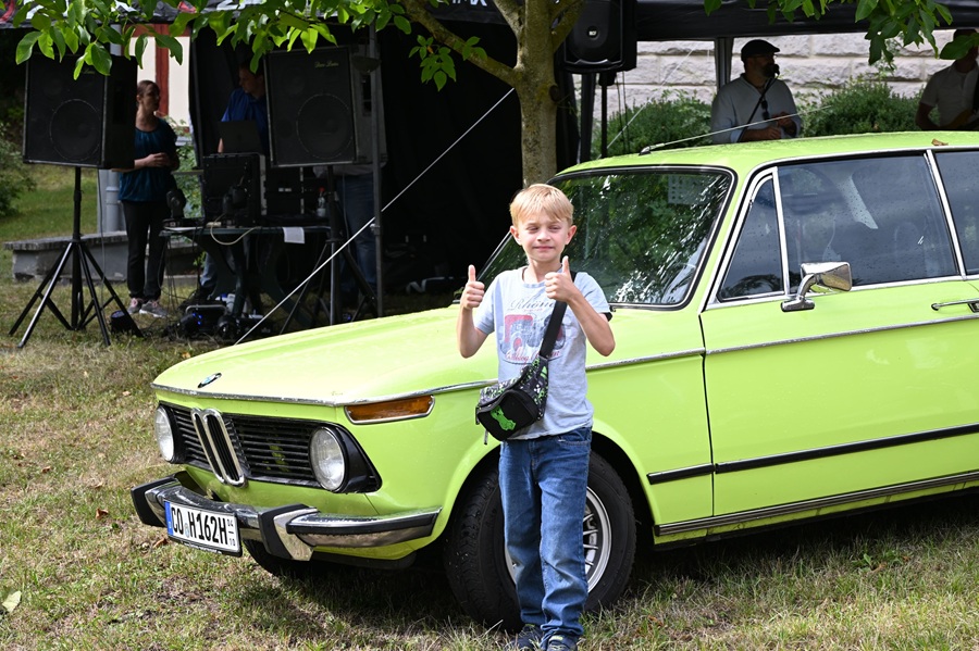 Ein laechelnder Junge zeigt Daumen hoch vor einem gruenen Oldtimer