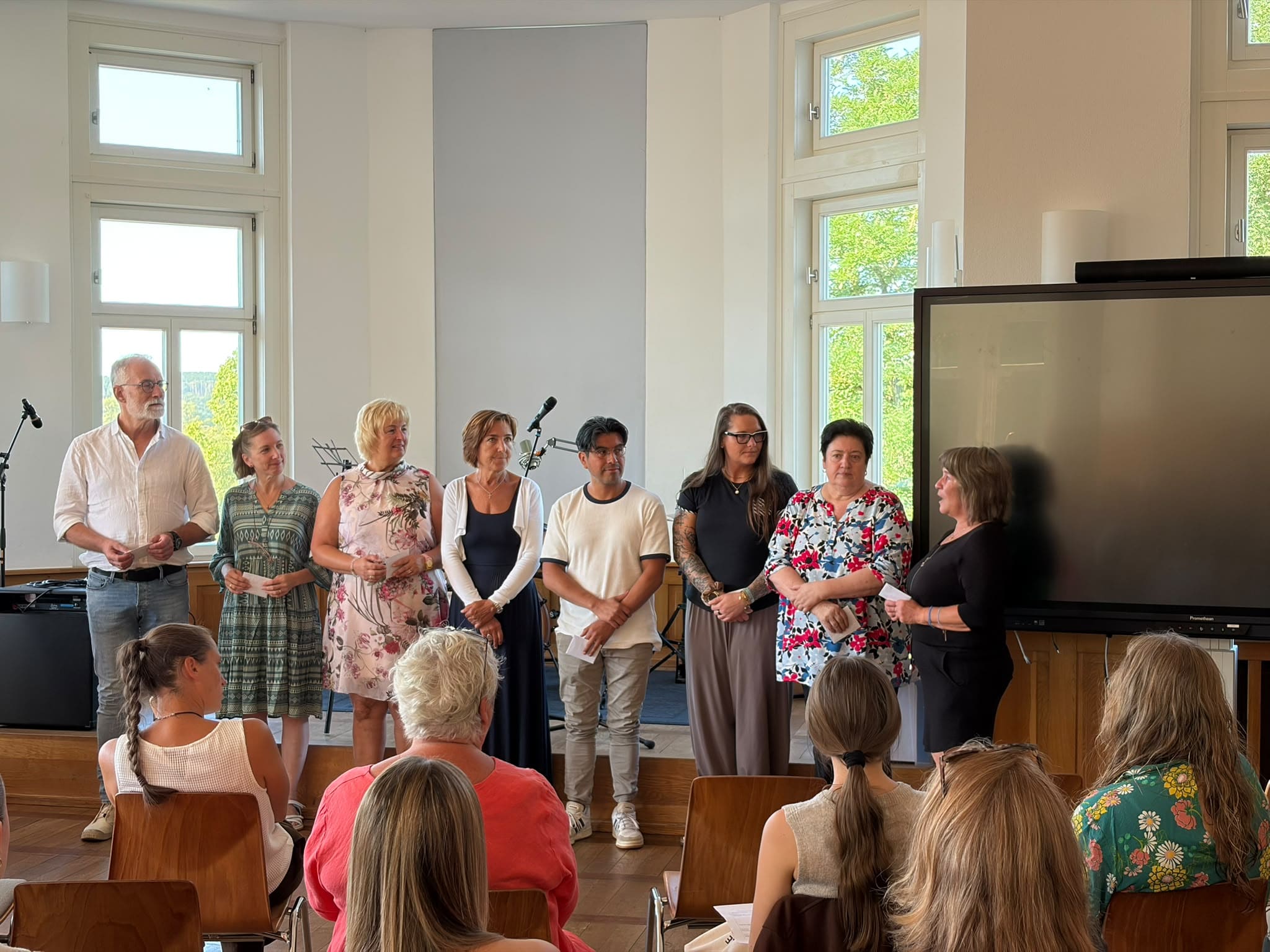 Eine Gruppe festlich gekleideter Personen steht laechelnd vor sitzendem Publikum