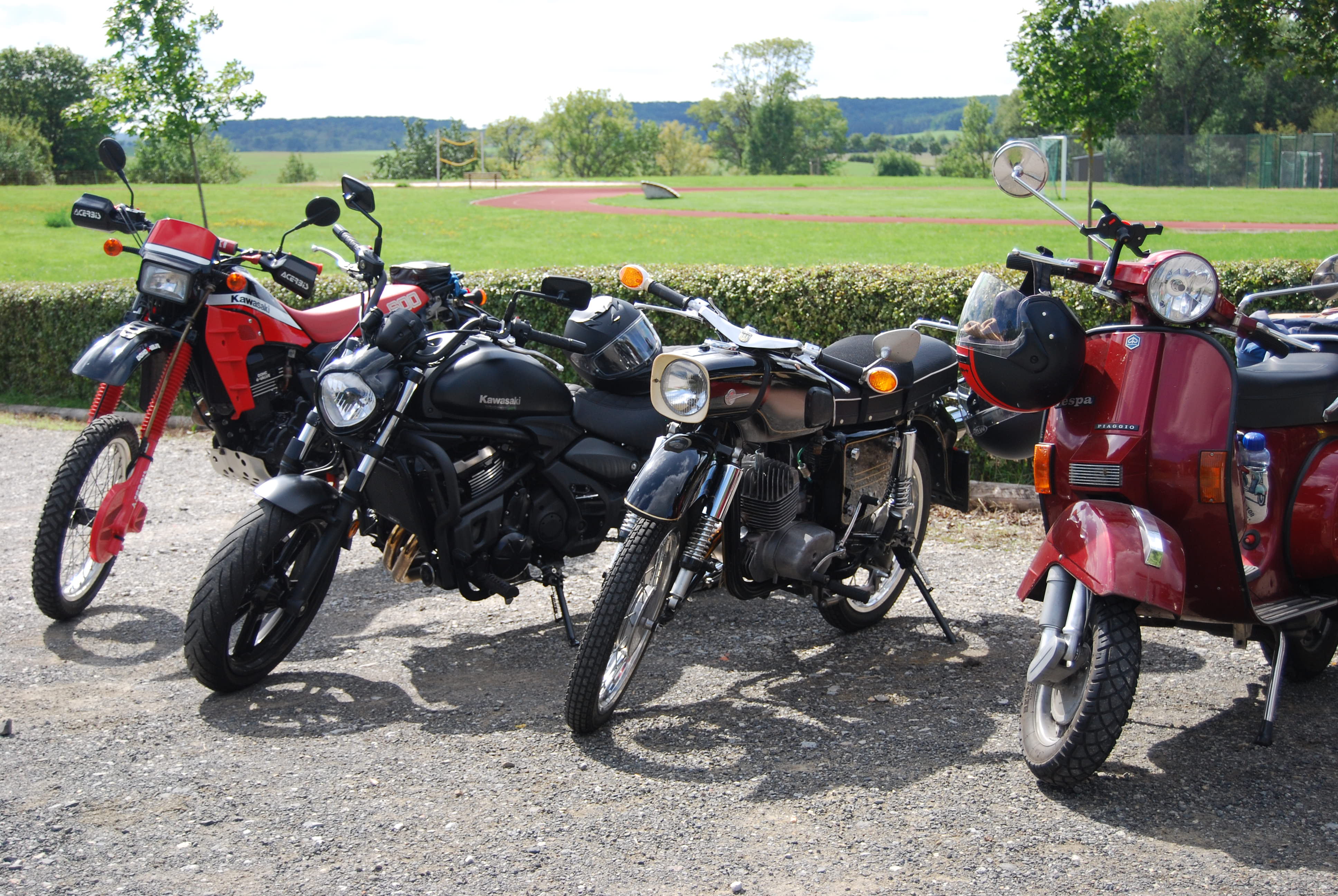 Verschiedene Motorraeder und eine rote Vespa stehen auf einem Kiesplatz