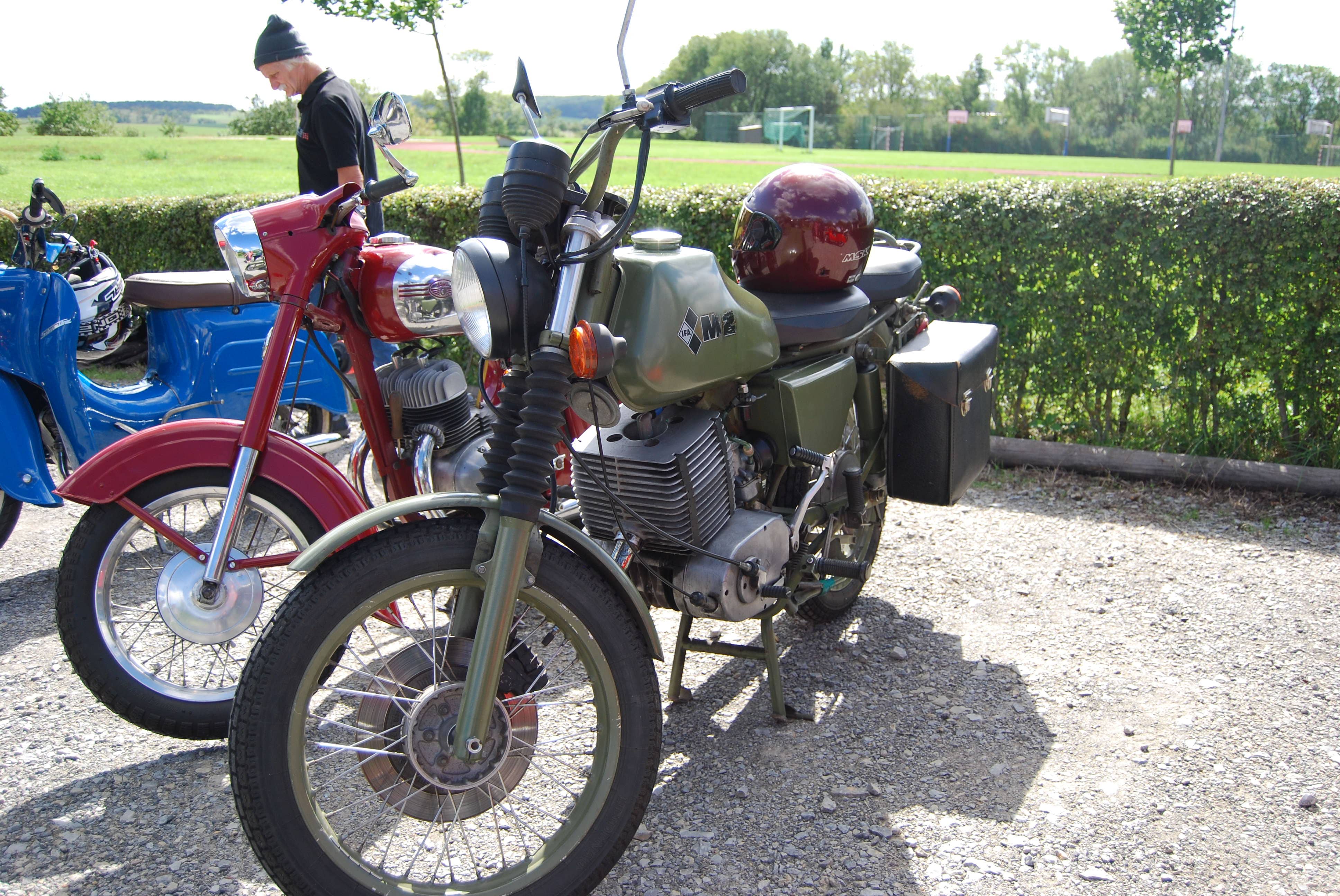 Olivgruenes Oldtimer Motorrad mit rotem Helm neben rotem und blauem Fahrzeug