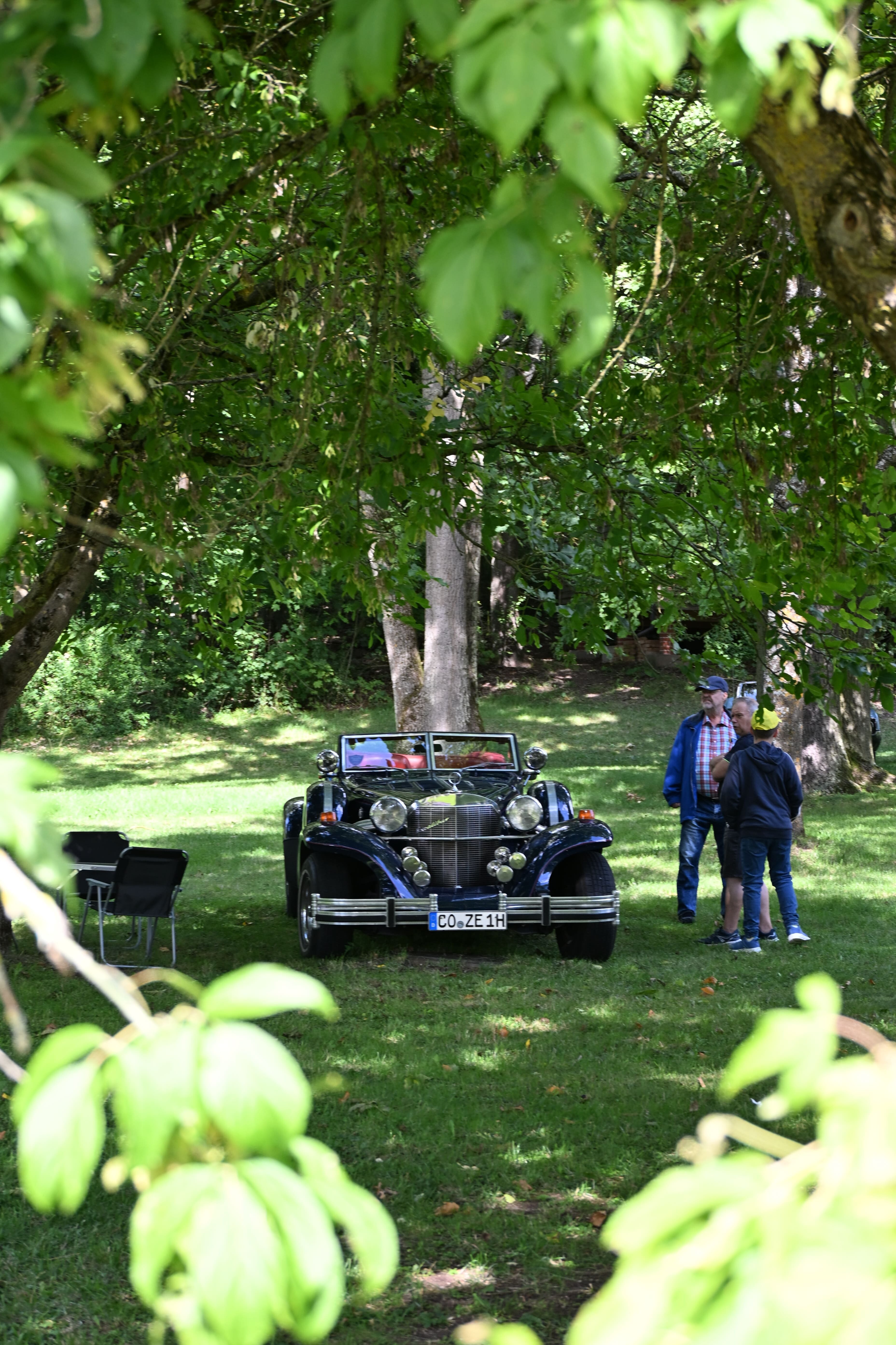 Dunkelblauer Oldtimer auf Wiese mit zwei Personen davor umgeben von Baeumen