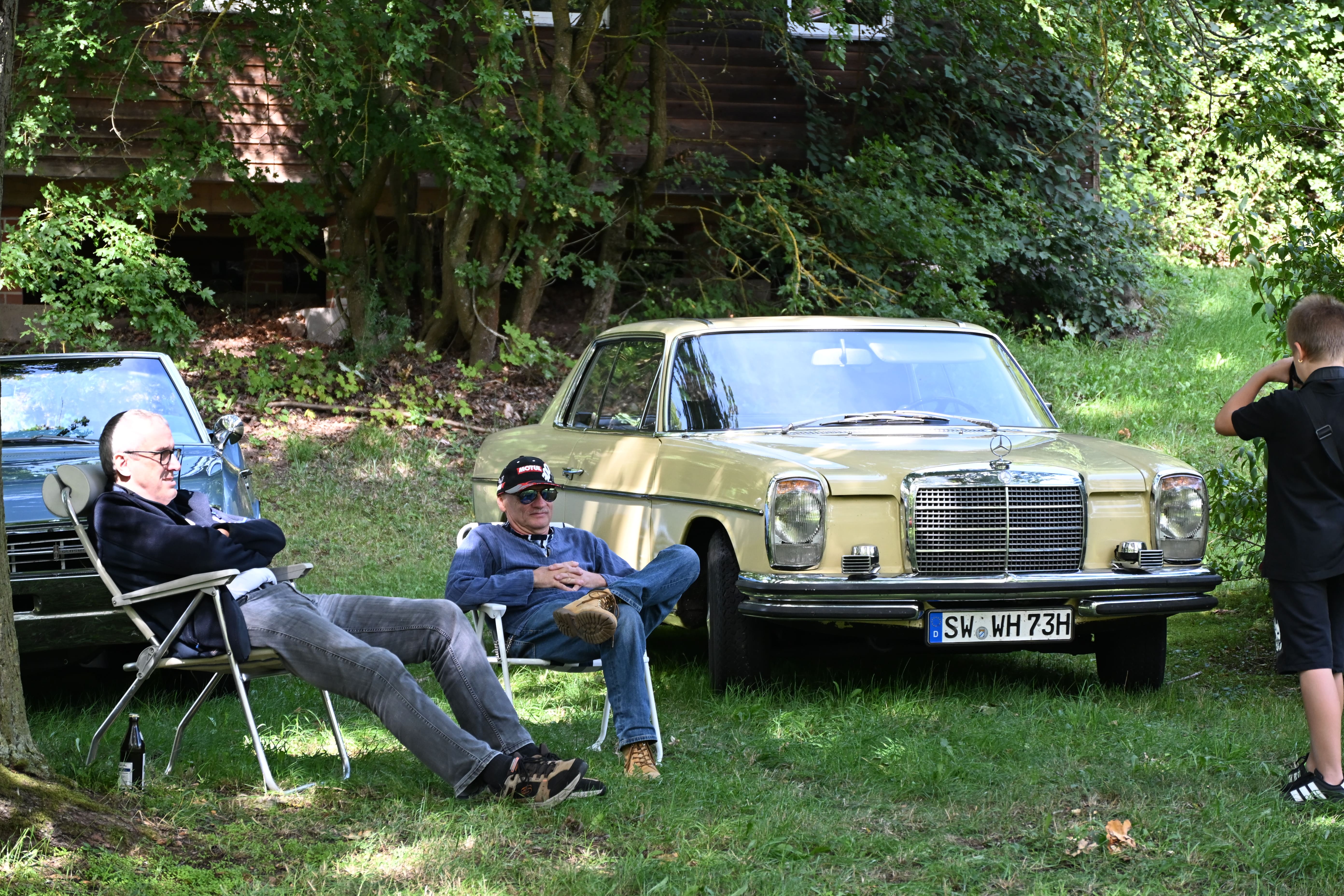 Zwei entspannte Maenner sitzen auf Campingstuehlen vor einem gelben Oldtimer