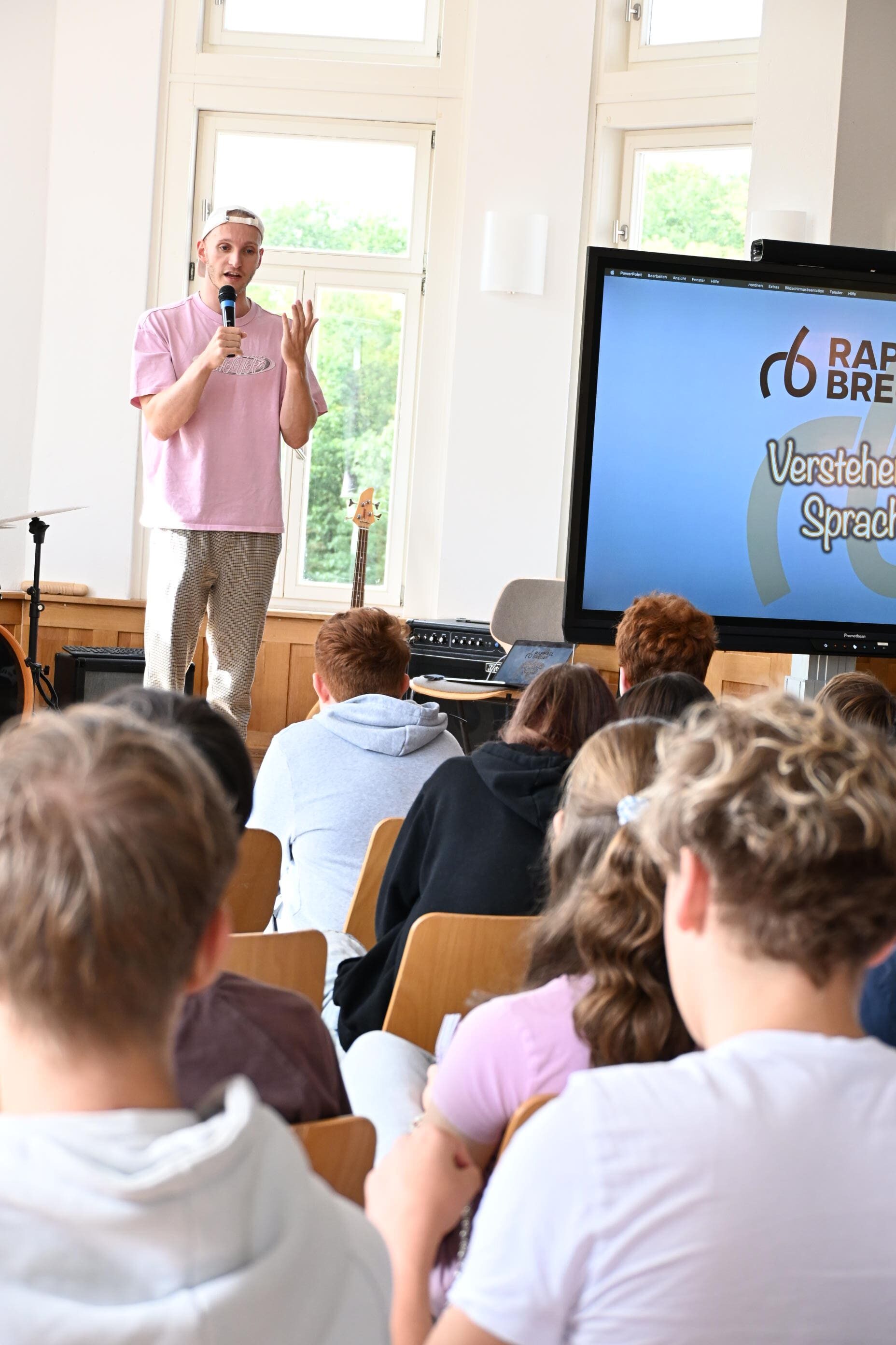 Ein junger Mann im rosa Shirt spricht mit Mikrofon vor Schuelern im Klassenraum