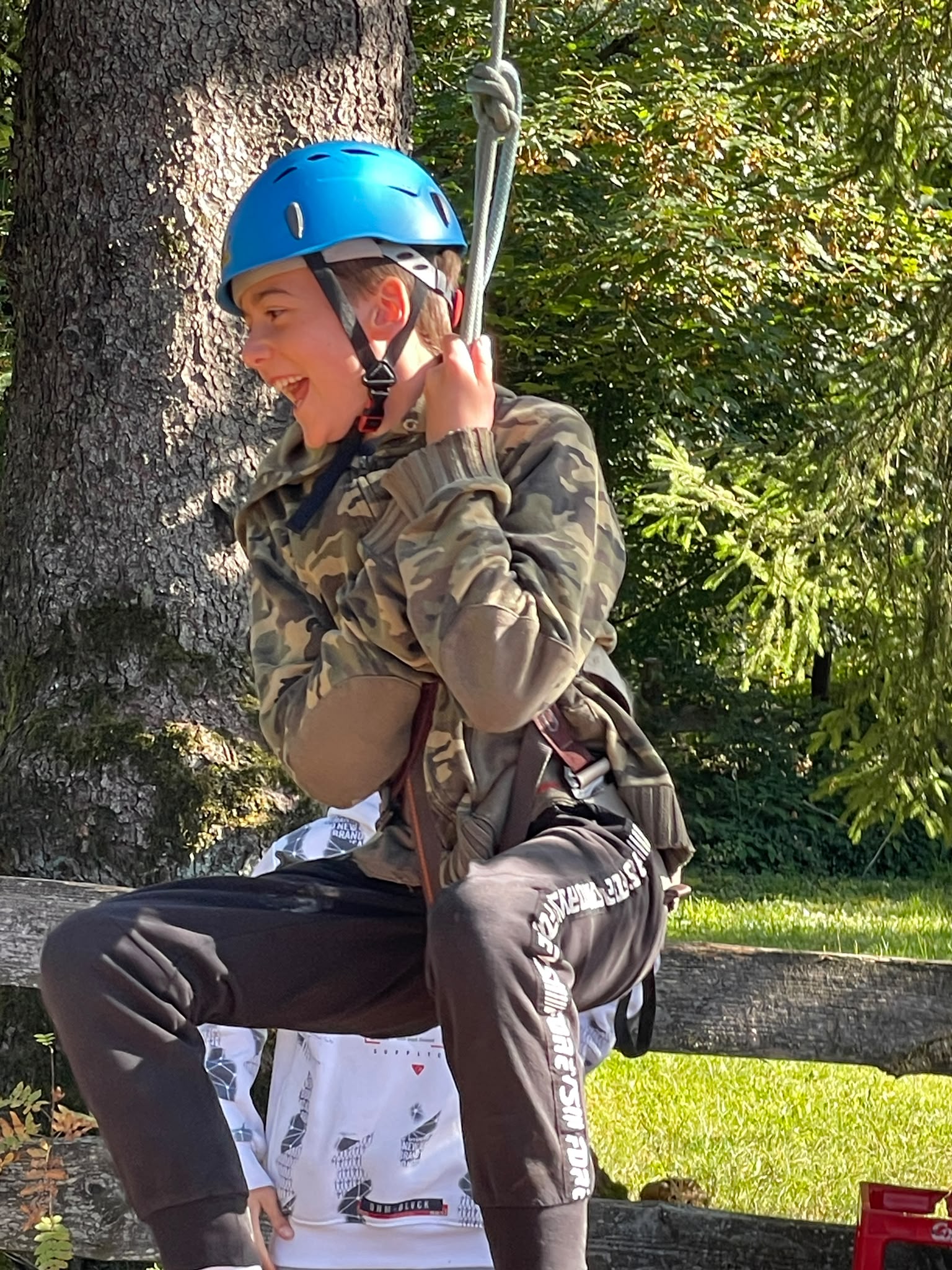 Ein lachender Junge mit blauem Helm und Tarnjacke klettert an einem Seil