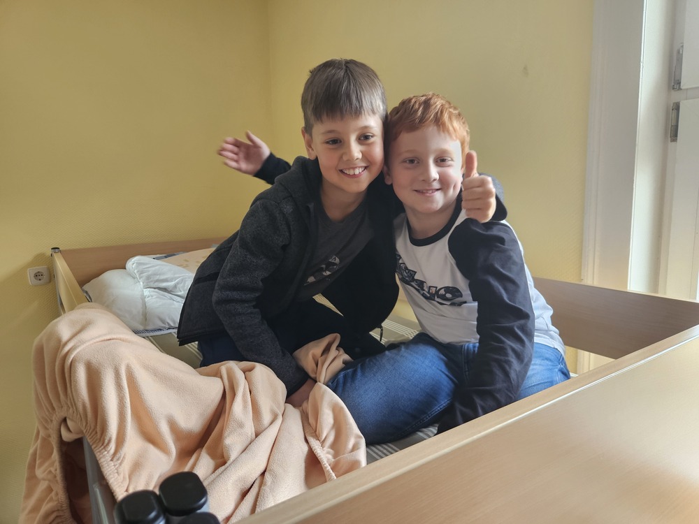 Zwei laechelnde Jungen sitzen umarmt auf einem Bett einer zeigt den Daumen hoch
