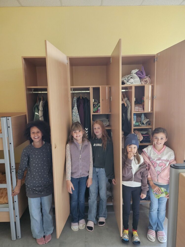 Fuenf laechelnde Kinder stehen vor offenen Holzschraenken in einem Zimmer