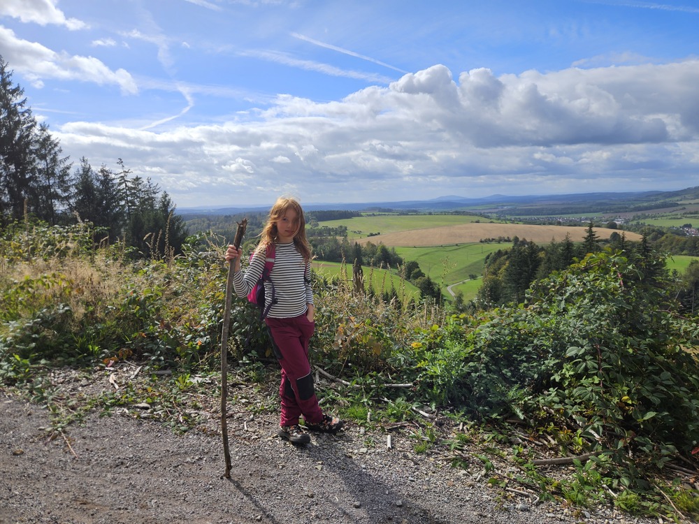 Ein Maedchen mit Wanderstock und Streifenshirt vor huegeliger Landschaft