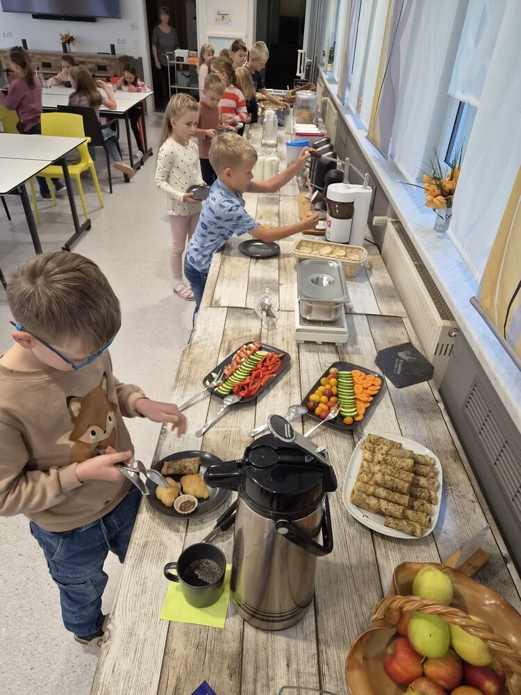 Kinder stehen an einem Buffet mit Gemuese Obst und Gebaeck und holen sich Essen
