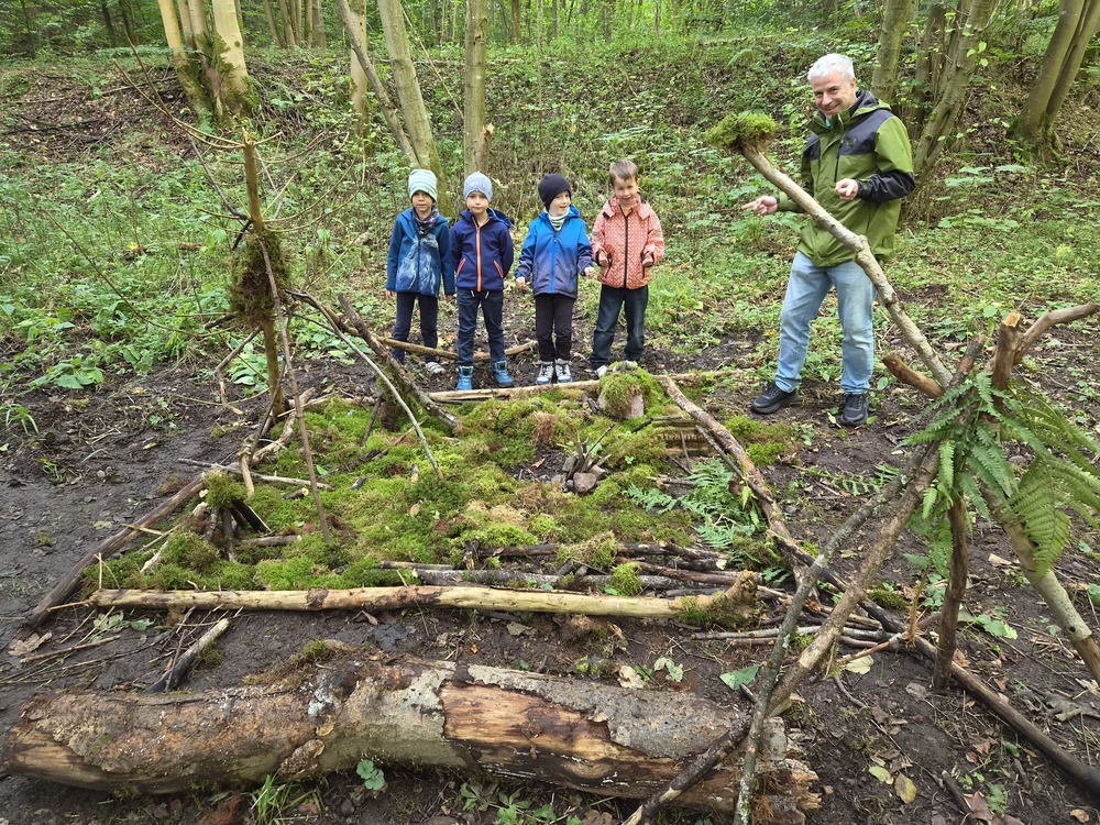 Erwachsener und vier Kinder bauen froehlich ein Waldlager aus Aesten und Moos