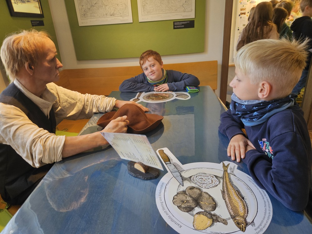 Mann in historischer Kleidung spricht mit zwei Jungen am blauen Tisch im Museum