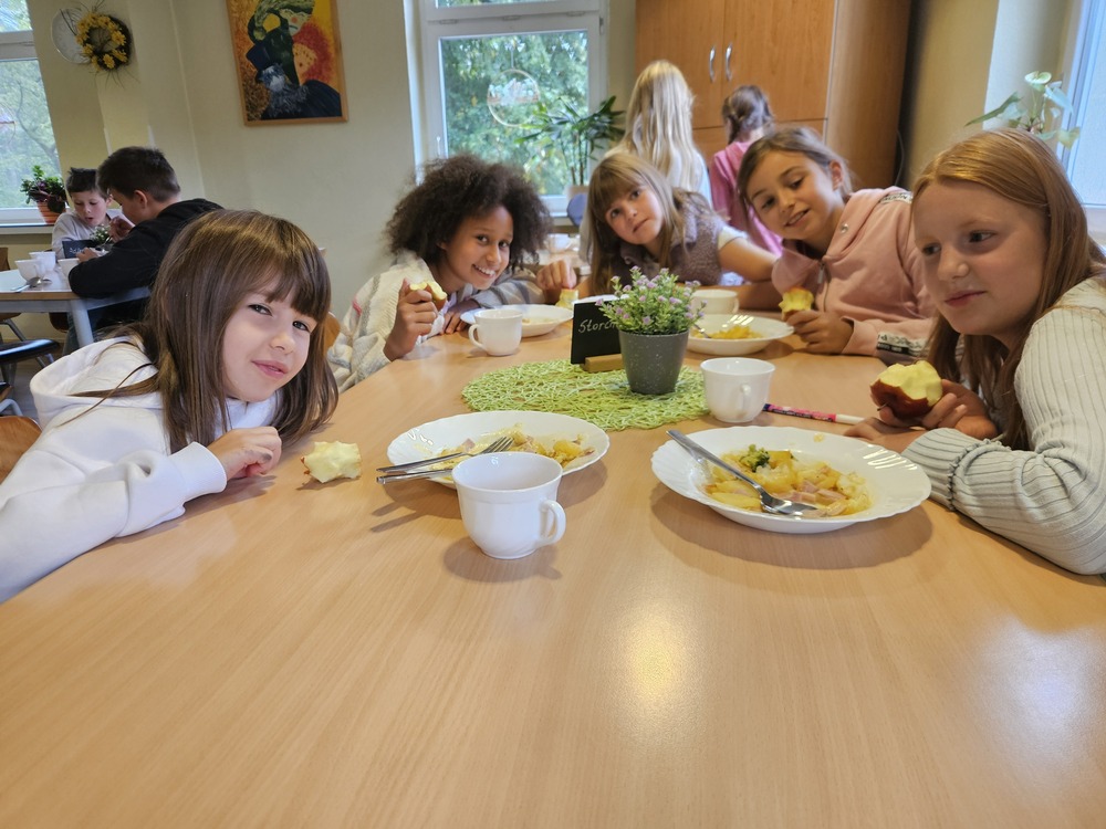 Laechelnde Kinder beim gemeinsamen Essen an einem Holztisch im Klassenzimmer