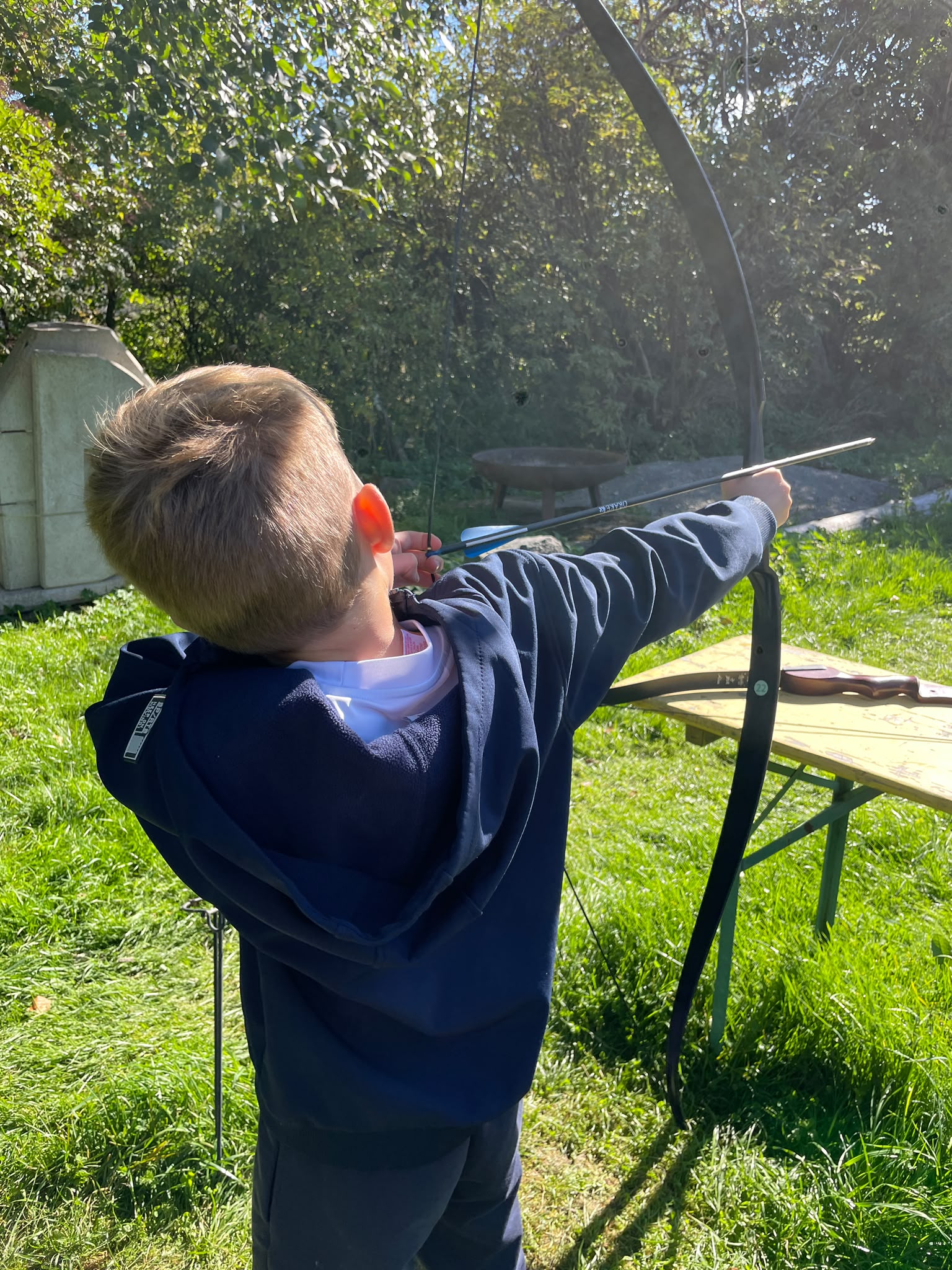 Konzentrierter blonder Junge im blauen Pullover zielt mit Pfeil und Bogen im Garten