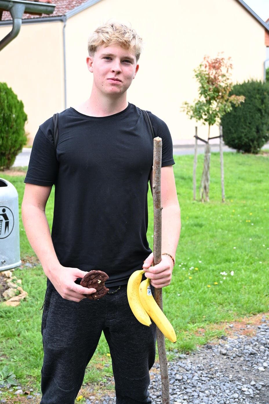 Blonder junger Mann in Schwarz haelt Bananen und Keks im Garten
