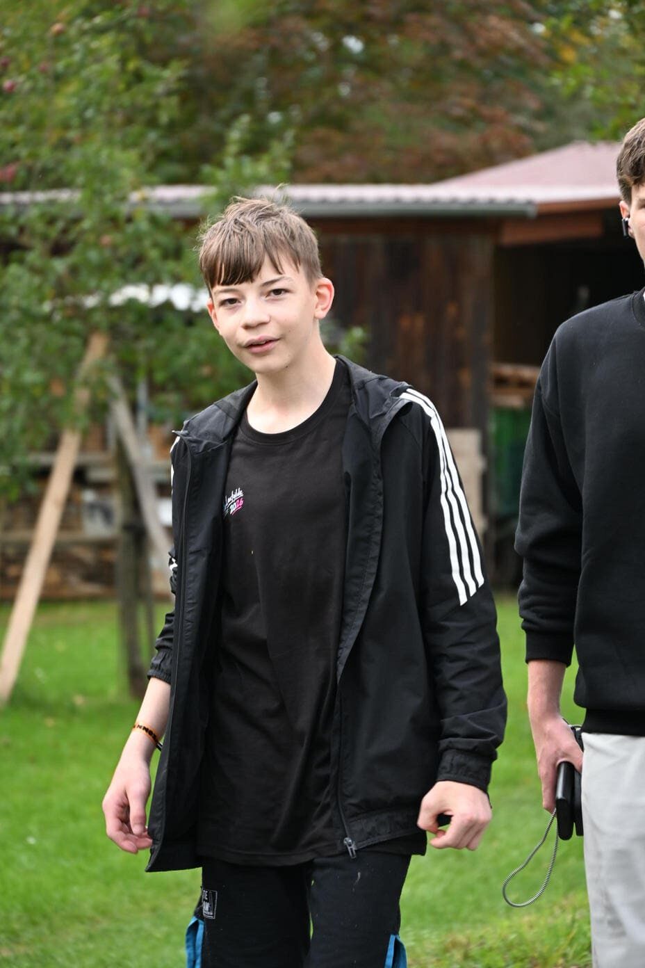 Ein Junge mit blonden Haaren traegt eine schwarze Jacke im Freien
