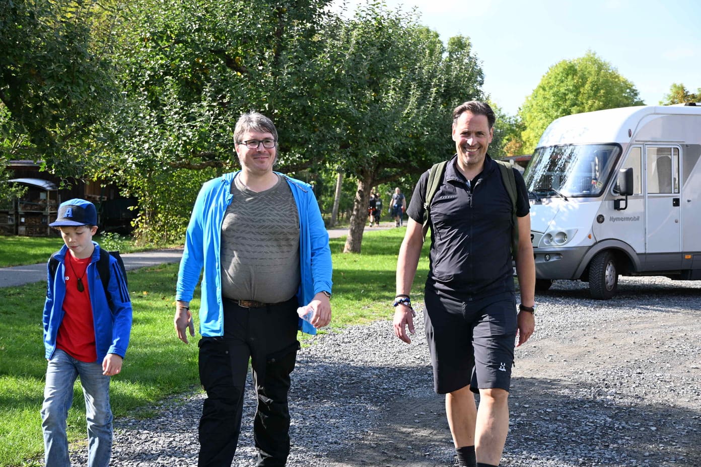 Zwei laechelnde Maenner und ein Junge gehen auf einem Kiesweg am Wohnmobil vorbei