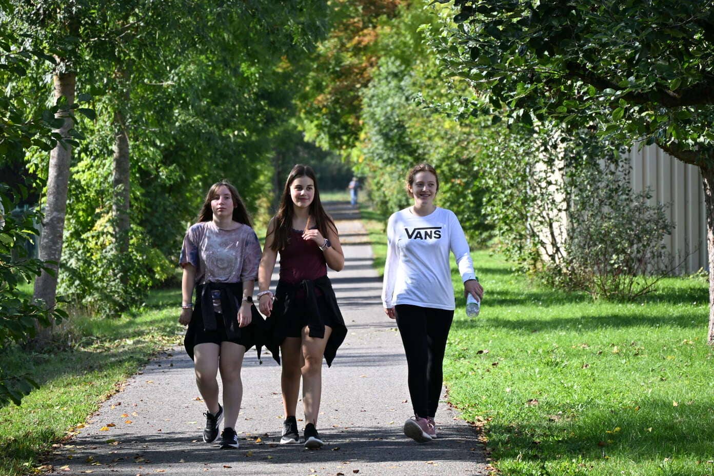 Drei laechelnde junge Frauen gehen auf einem von Baeumen gesaeumten Weg
