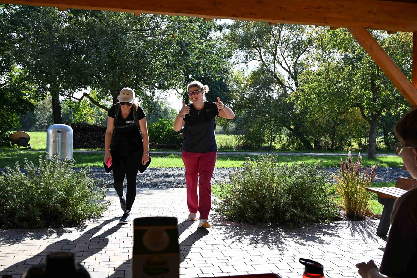 Zwei laechelnde Frauen gehen durch einen Park, eine zeigt beide Daumen hoch