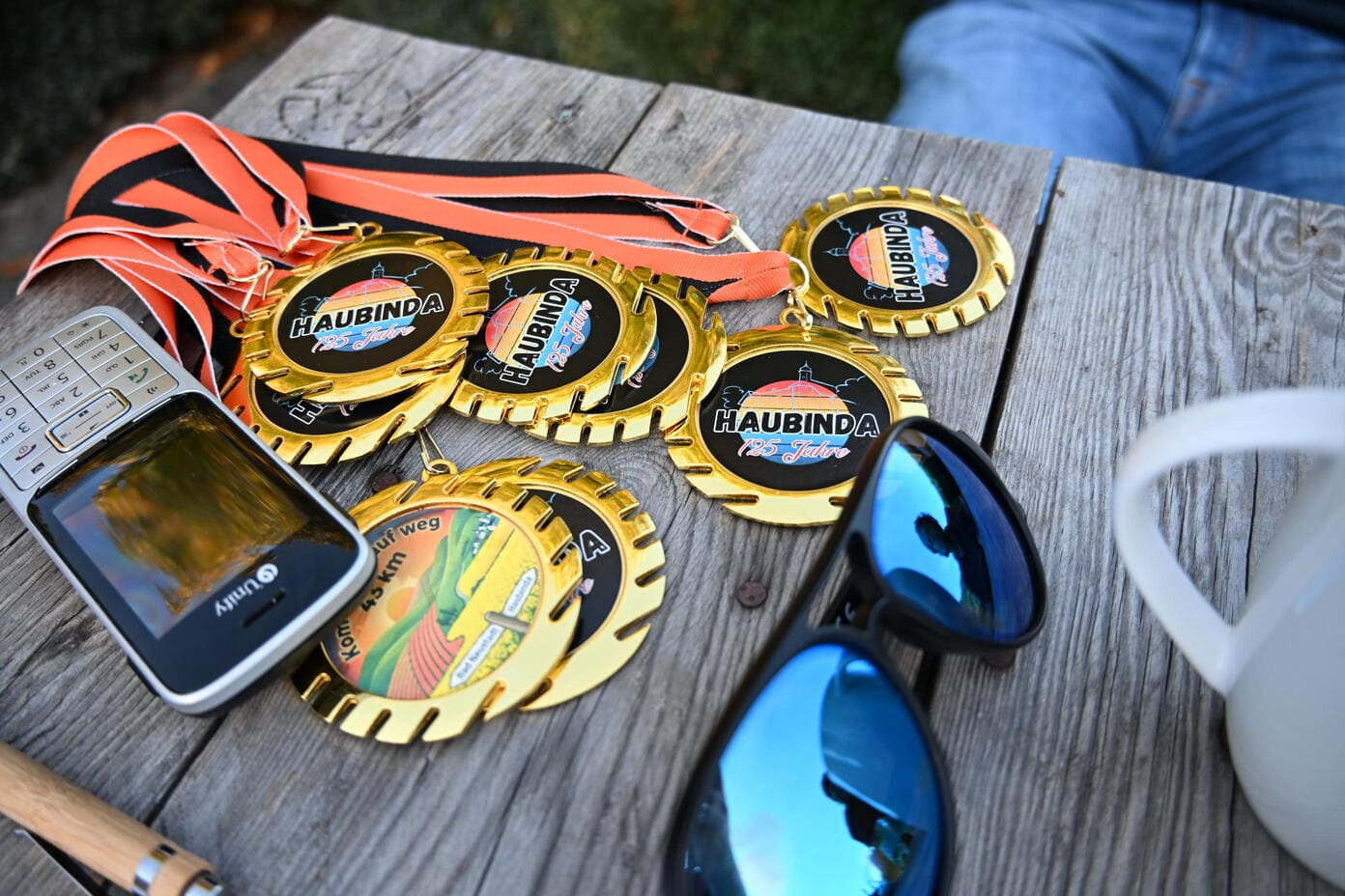 Goldene Haubinda Medaillen mit orangen Baendern neben Handy und Sonnenbrille