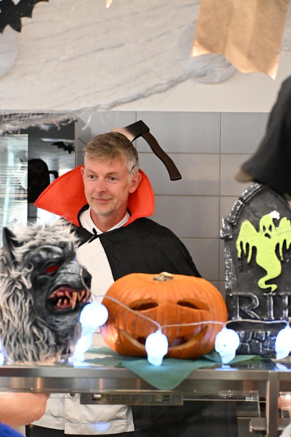 Laechelnder Mann im Vampirkostuem mit Beilstirnband hinter Halloween Deko