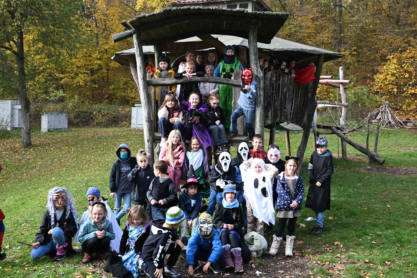 Eine Gruppe Kinder in bunten Halloweenkostuemen posiert an einem Spielhaus im Wald