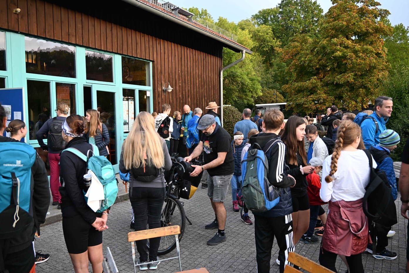 Eine Gruppe von Menschen mit Rucksaecken versammelt sich vor einem Holzgebaeude