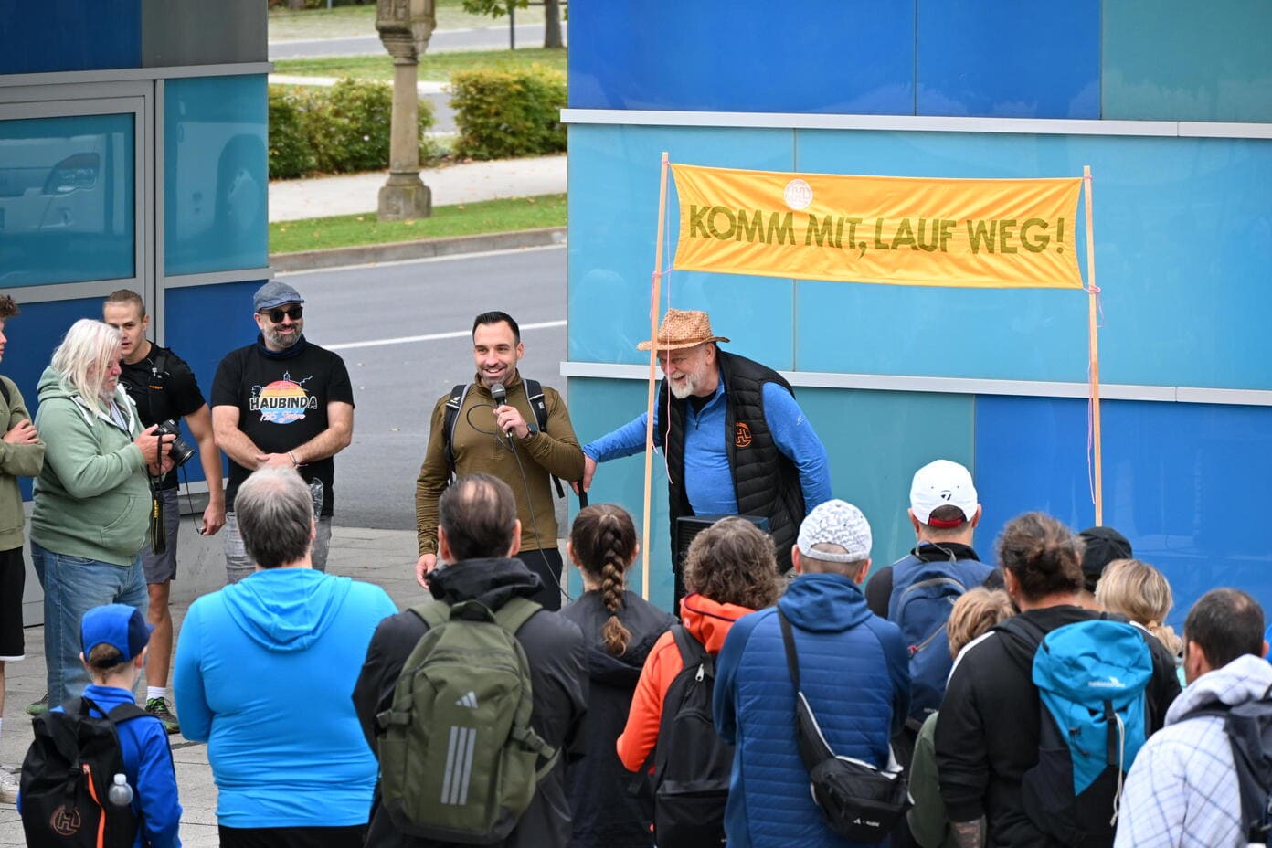 Ein laechelnder Mann mit Mikrofon spricht vor Menschen unter einem gelben Banner