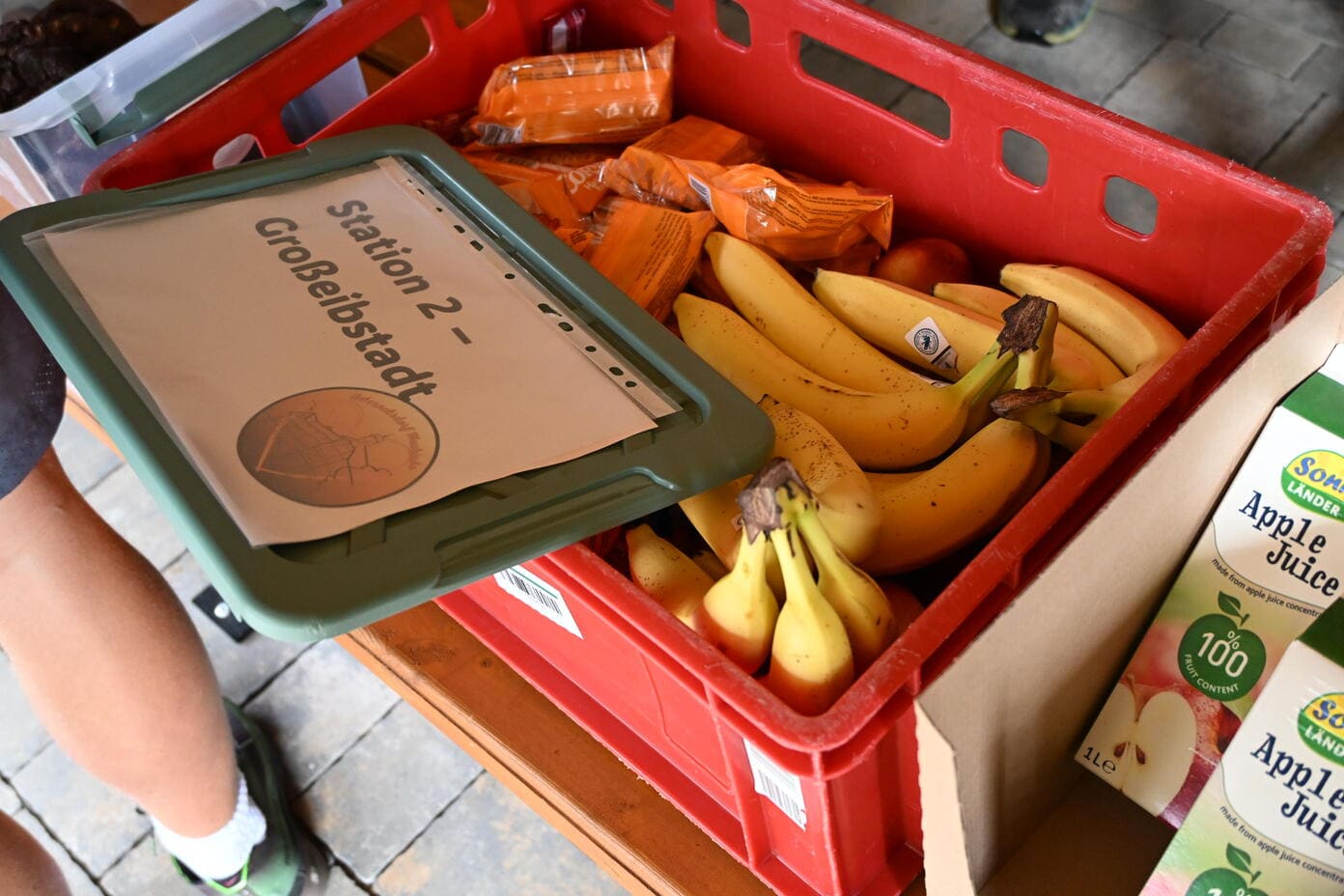 Rote Kiste mit gelben Bananen und orangen Packungen neben Apfelsaftkartons
