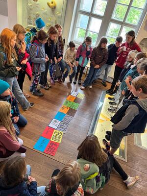 Kinder stehen und sitzen um bunte Karten auf dem Boden in einem Museumsraum