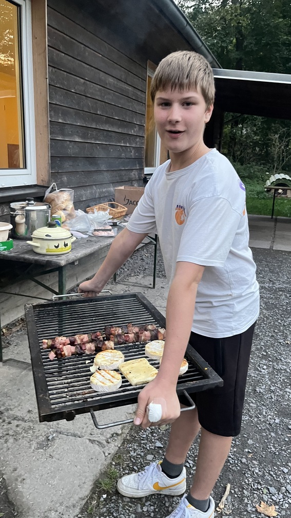 Junger Mann im weissen Shirt haelt laechelnd einen Grill mit Fleisch und Kaese