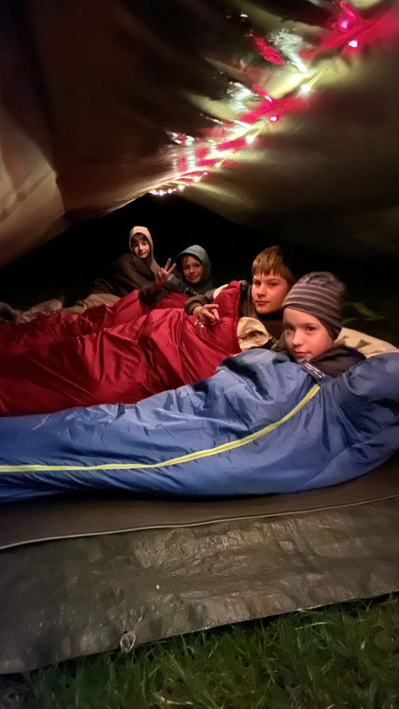 Vier laechelnde Kinder in blauen und roten Schlafsaecken in einem Zelt bei Nacht