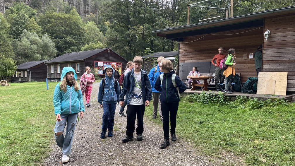 Kinder in bunten Jacken gehen einen Weg entlang an Holzhuetten im Wald
