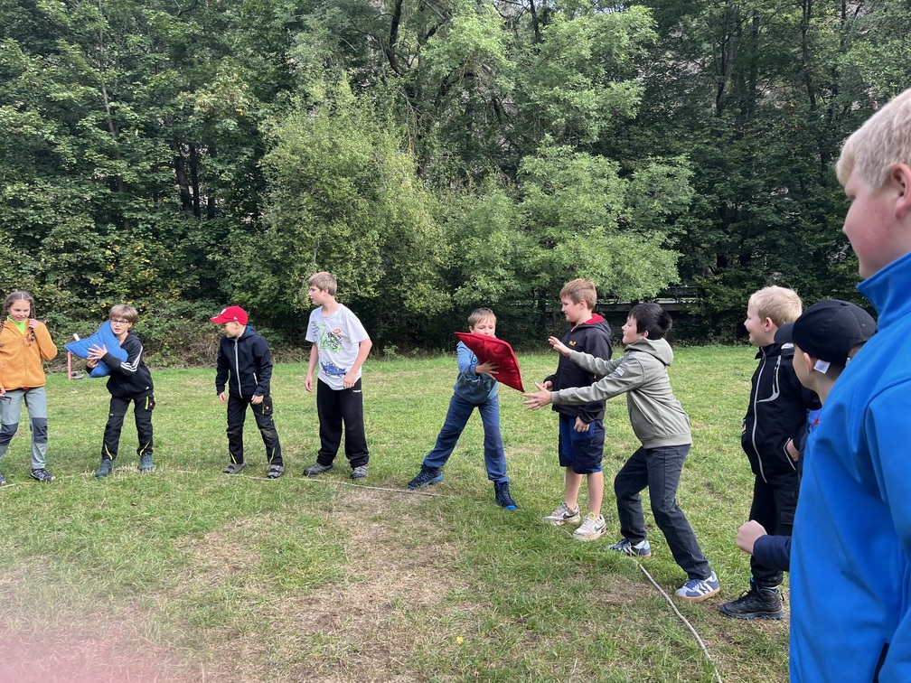 Kinder spielen konzentriert auf einer Wiese vor gruenen Baeumen mit bunten Kissen