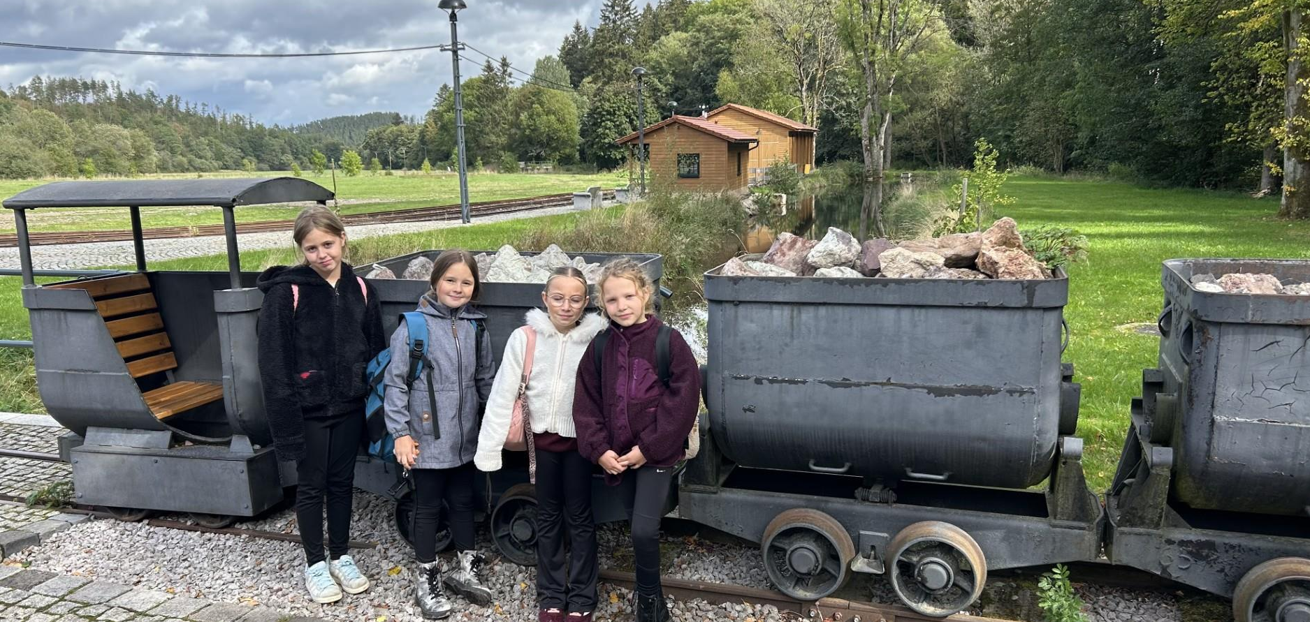 Vier laechelnde Maedchen stehen vor alten Grubenwagen mit Steinen in Natur
