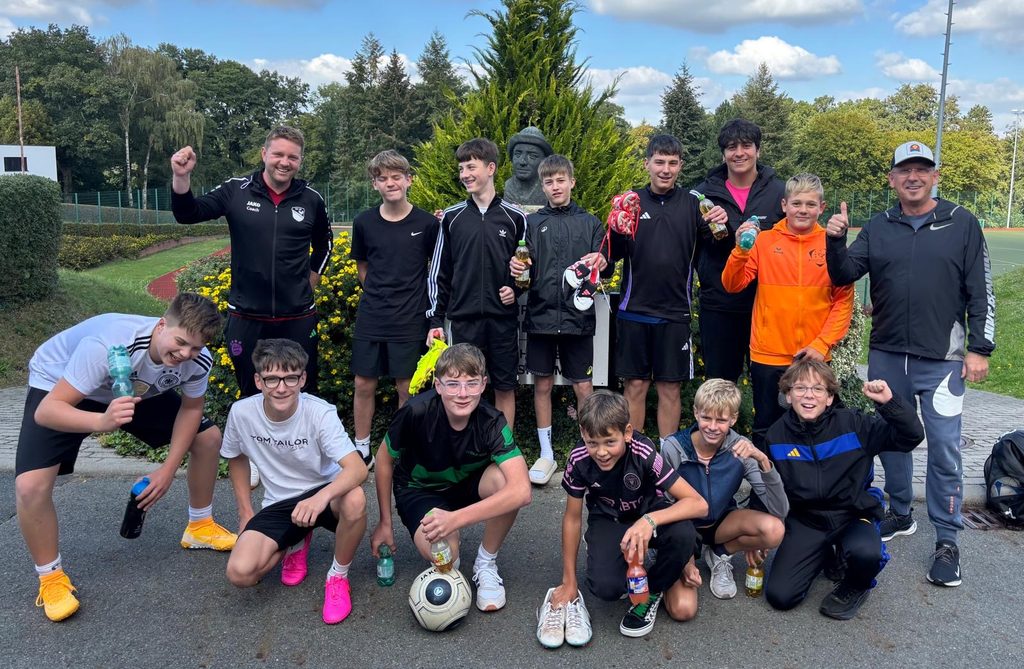 Froehliche Jugendfussballmannschaft mit zwei Trainern posiert vor einem Sportplatz